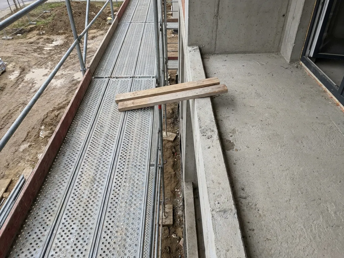 Pohled šikmo shora na mezeru mezi podlahou fasádního lešení a betonovým balkonem, přes kterou leží dvě krátké desky.